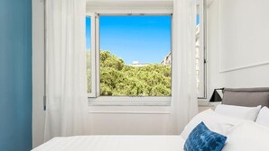Un dormitorio con un ventanal grande, cortinas blancas, una cama con almohadas blancas y azules, y vistas a zonas verdes y edificios.