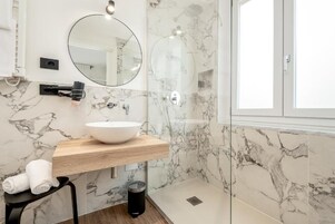 A modern bathroom with a marble backsplash, a wooden vanity, a round mirror, and a glass-enclosed shower.