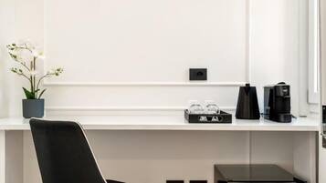A modern, minimalist room with a white wall, a black chair, a small potted plant, a coffee maker, and a black appliance on the floor.