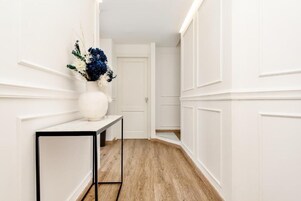 Valoisa käytävä, jonka seinät ovat valkoiset, lattia on puuta ja käytävän päässä on valkoinen ovi.