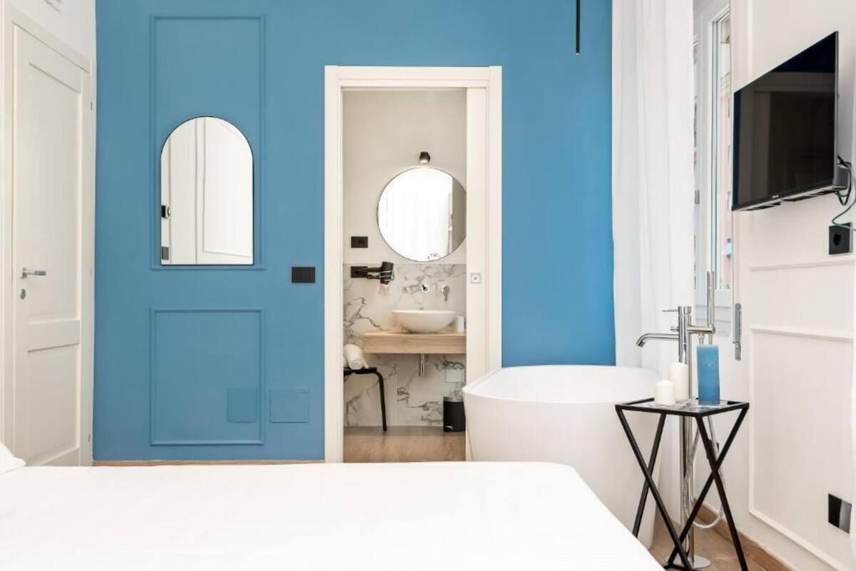 Ein modernes Badezimmer mit blauer Akzentwand, einer weißen Badewanne, einem runden Spiegel und einem angebrachten Fernseher.
