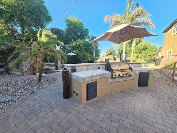 An outdoor barbecue area with a stone grill, a large umbrella, and a paved patio. - Second Story Suite w/ Balcony (Queen Creek)