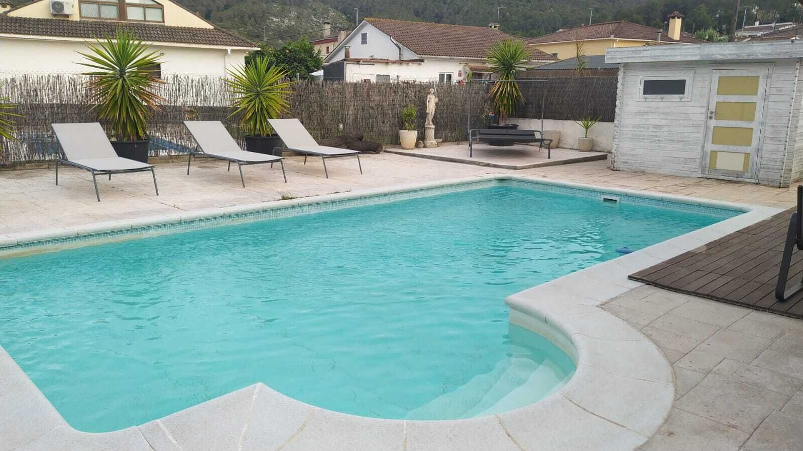 Una piscina rectangular amb vora corba, envoltada per un solàrium amb rajoles. Hi ha dues gandules i diverses plantes en testos.