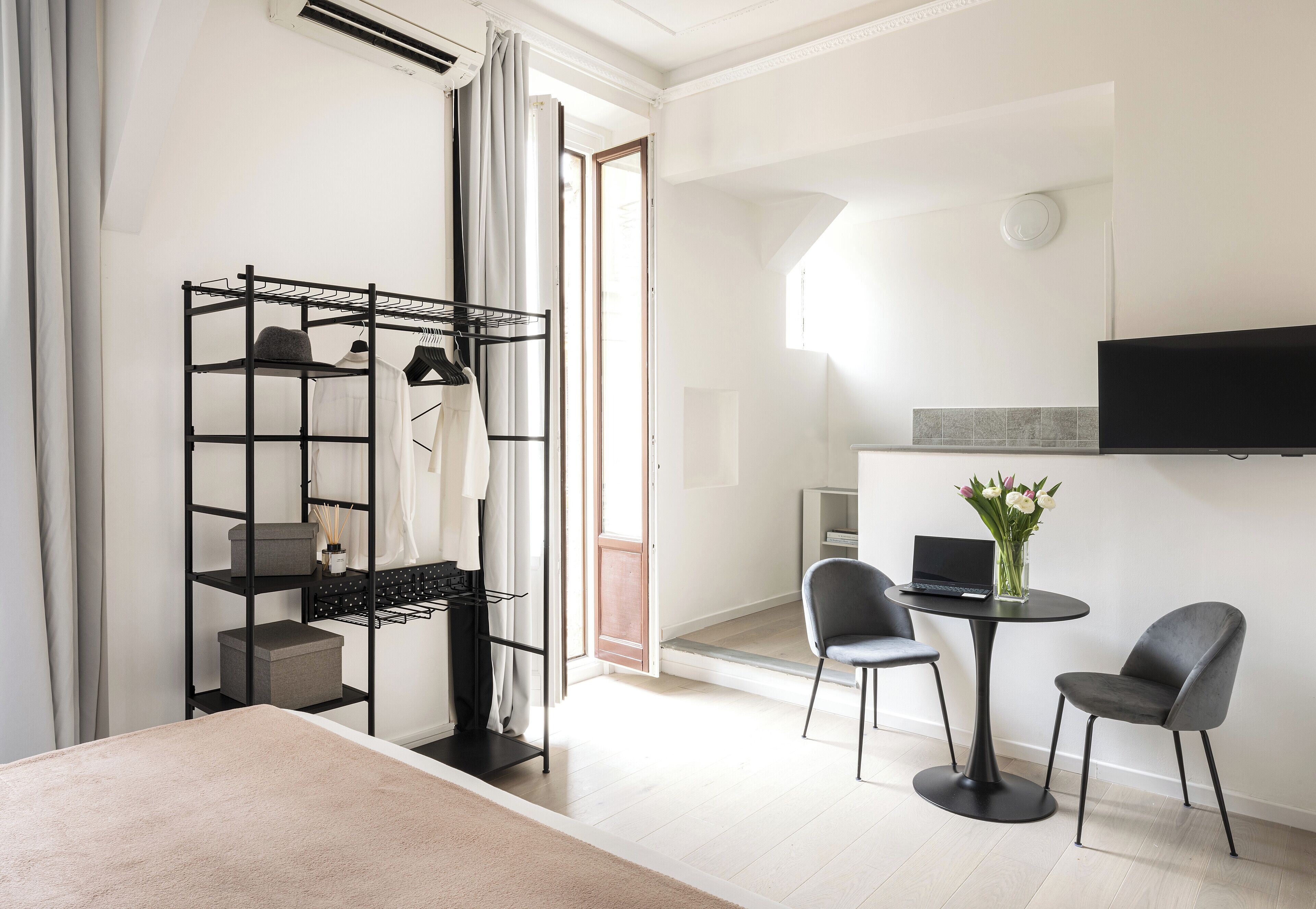 A modern, well-lit room with a black metal shelving unit, a dining table with two chairs, a TV mounted on the wall, and a large window.