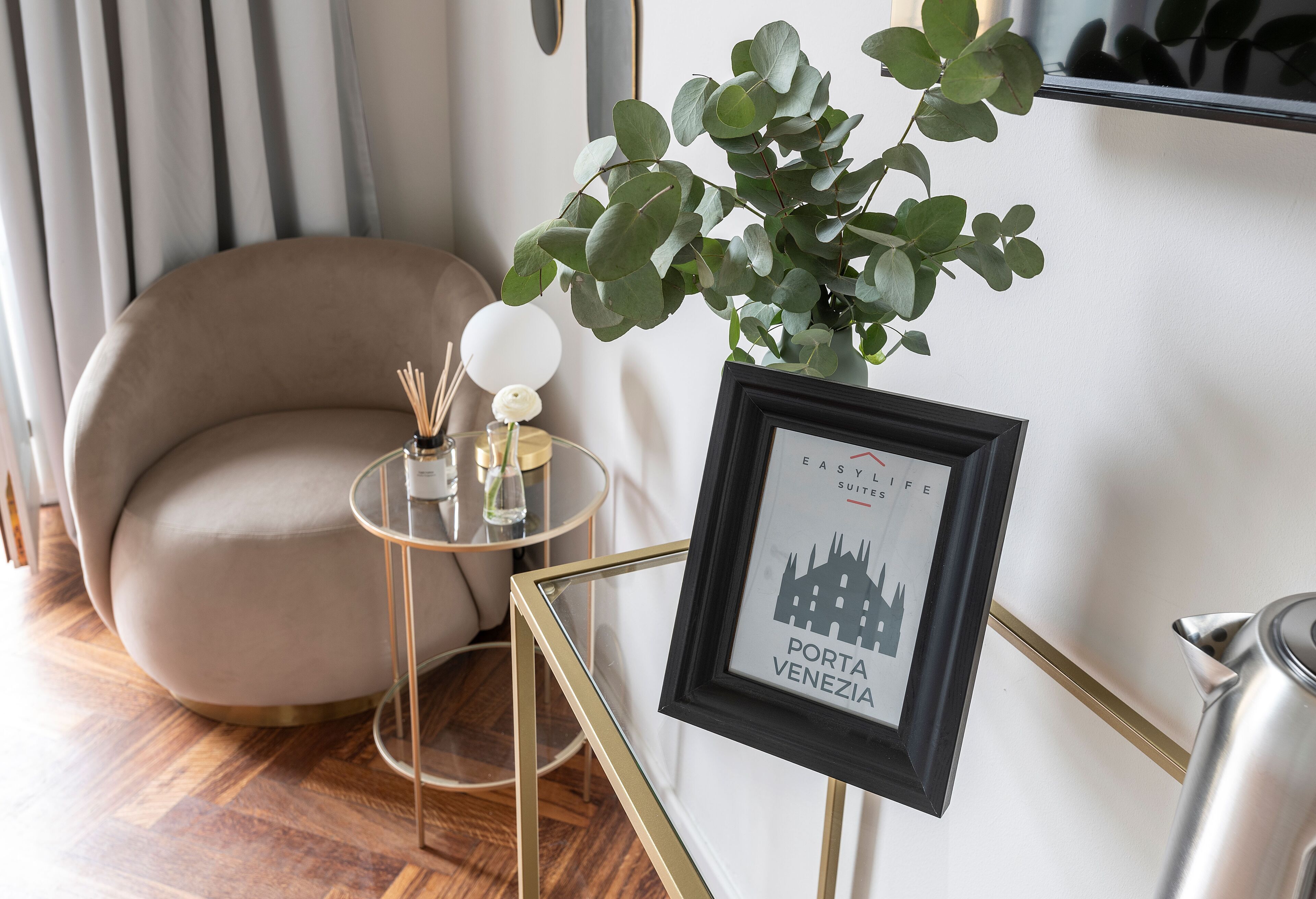 Un cuadro enmarco con una vista urbana, una mesa redonda con cubierta de vidrio, una silla color beige y una planta en maceta.