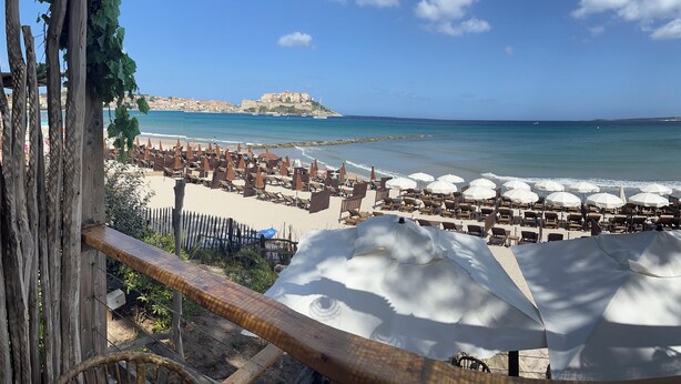 Une plage avec vue sur la mer, agrémentée de parasols et de chaises longues, une clôture en bois et, au loin, une structure qui ressemble à un château.