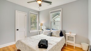 Una camera da letto con un letto, una ventola a soffitto, un comodino, una lampada e una finestra con vista su una casa.
