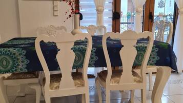 Un coin repas avec une table dressée pour deux personnes, des chaises blanches, une nappe à motifs, un vase avec des fleurs rouges et un tableau accroché au mur.