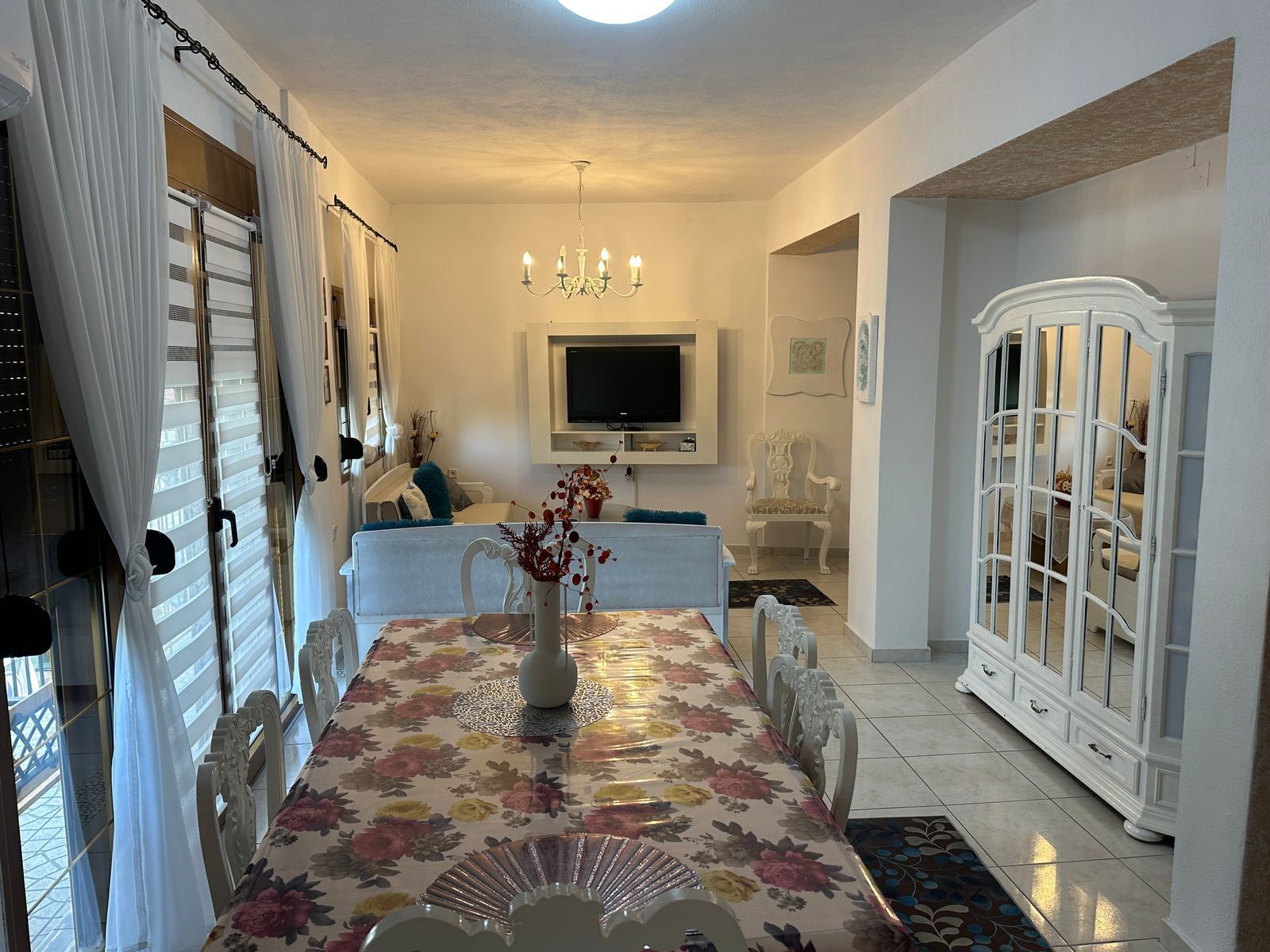 Un comedor con mantel floreado, una araña y un aparador blanco con puertas de vidrio.