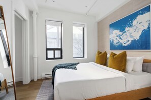 A bedroom with a large bed, two windows, a painting, and a wooden floor.
