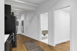 Una cocina moderna con un refrigerador negro, una estufa y un lavamanos. El suelo es de madera y hay una puerta blanca que da a un baño con un lavamanos y un espejo.