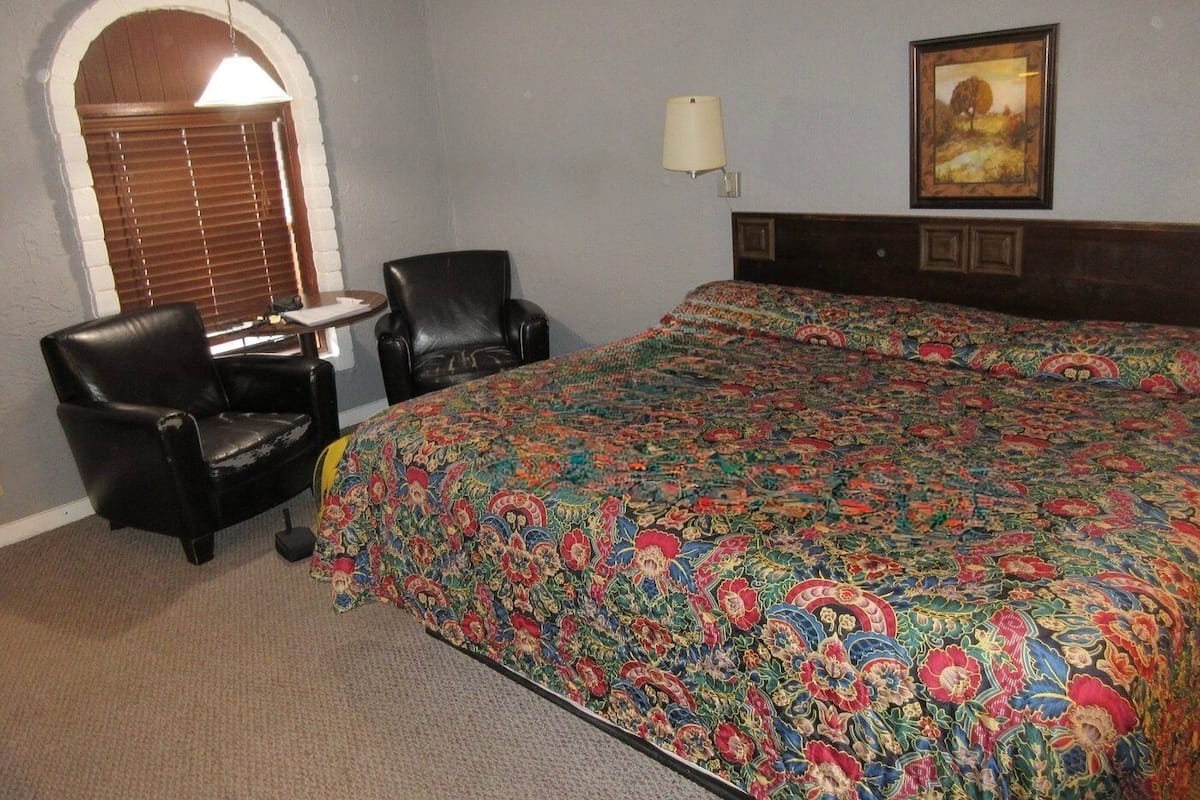Une chambre d’hôtel avec un grand lit, deux chaises, une petite table, une lampe et un tableau au mur.