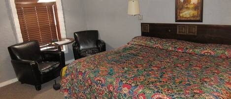 A hotel room with a large bed, two chairs, a small table, a lamp, and a painting on the wall.