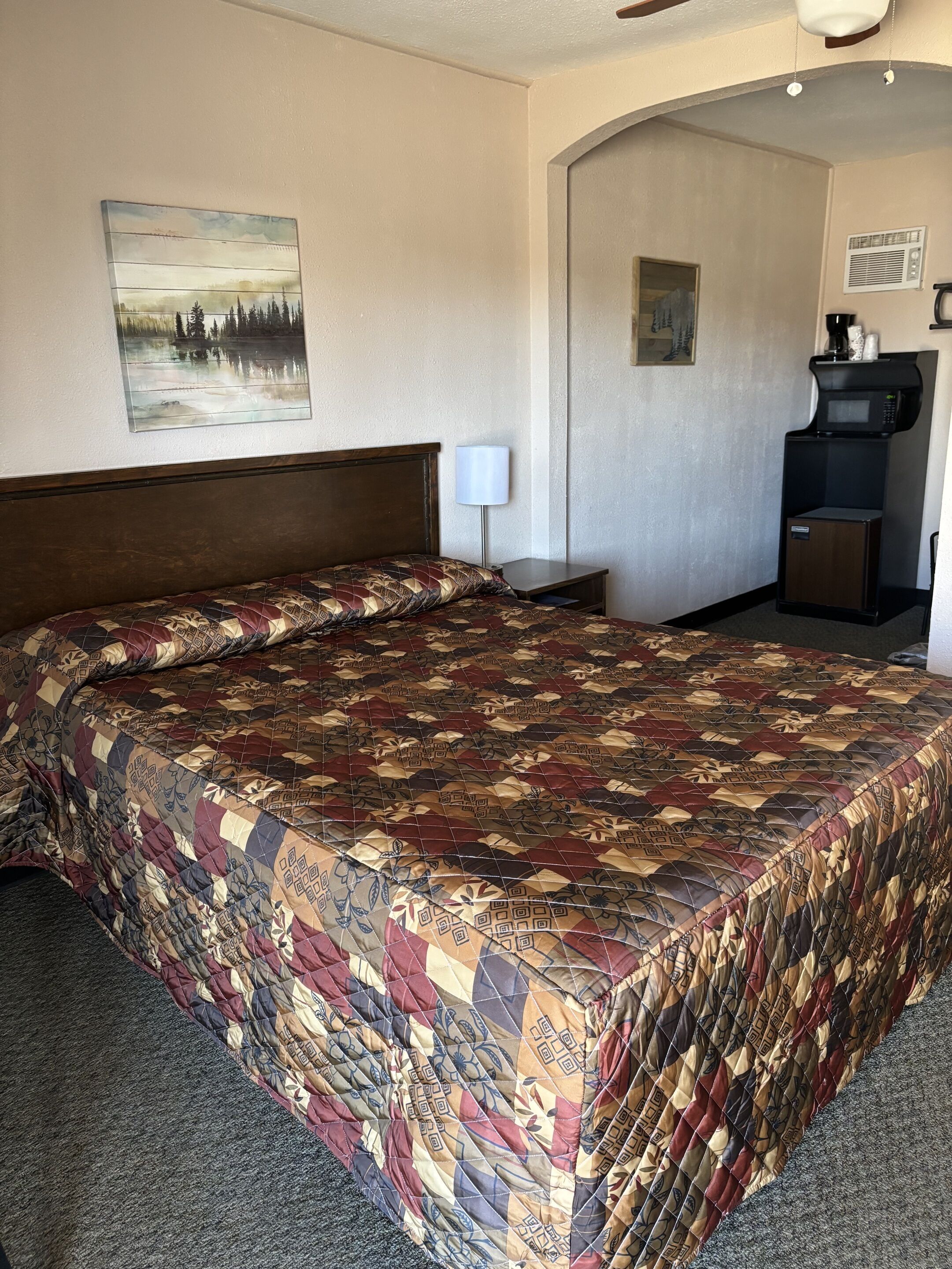 A hotel room with a large bed, a nightstand, a lamp, a painting of a lake, and a coffee machine.