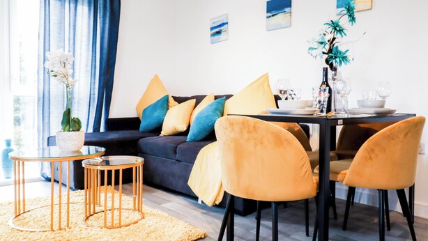 Ein modernes Wohnzimmer mit einem dunklen Sofa, gelben Kissen, einem Glastisch und einem Essbereich mit Tisch und Stühlen.