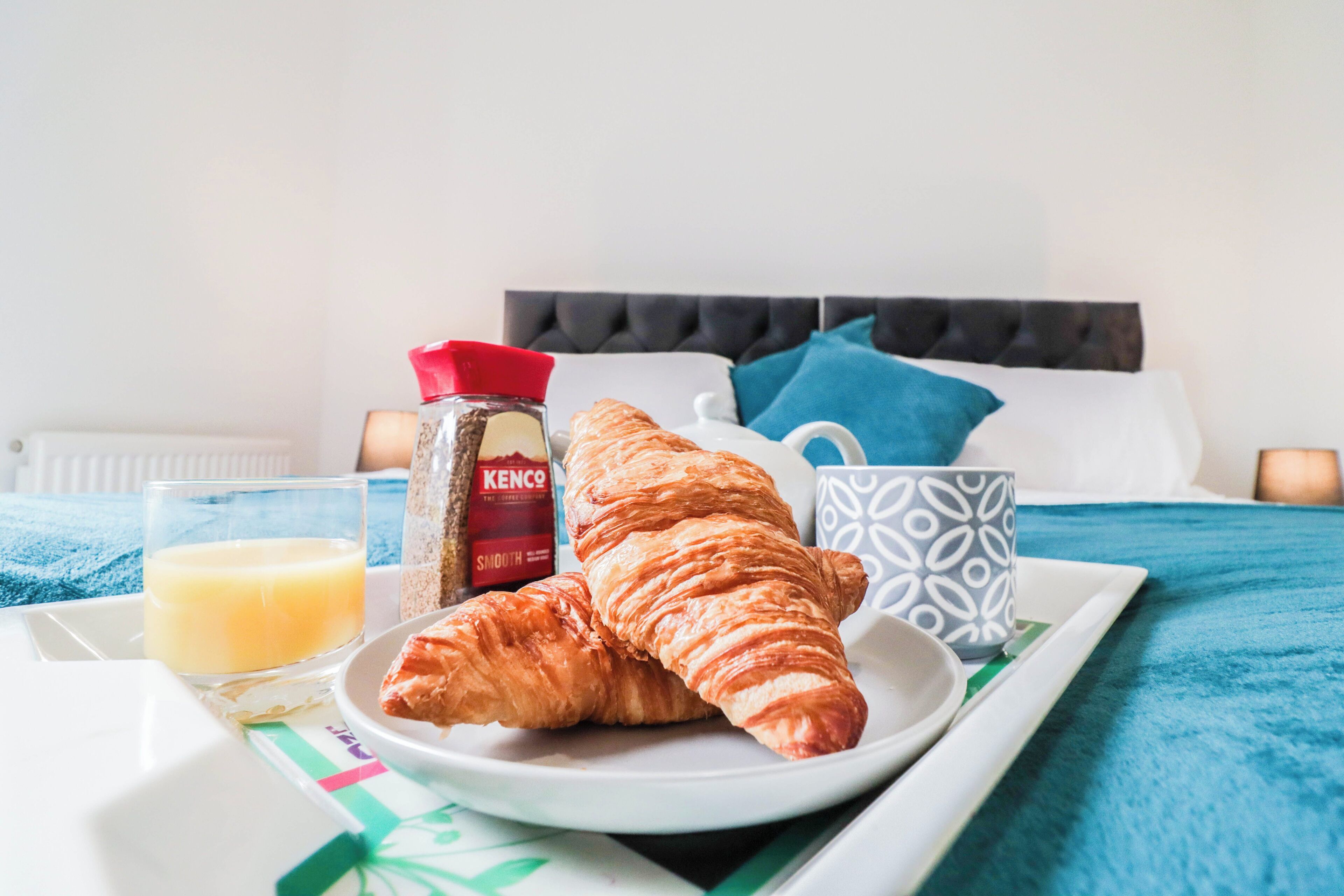 A breakfast tray with croissants, a glass of orange juice, a jar of Kenco coffee, and a patterned mug on a bed with a blue and white bedspread.