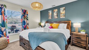 A bedroom with a large bed, a wooden nightstand, a wicker lamp, and a view of the outdoors through the window.