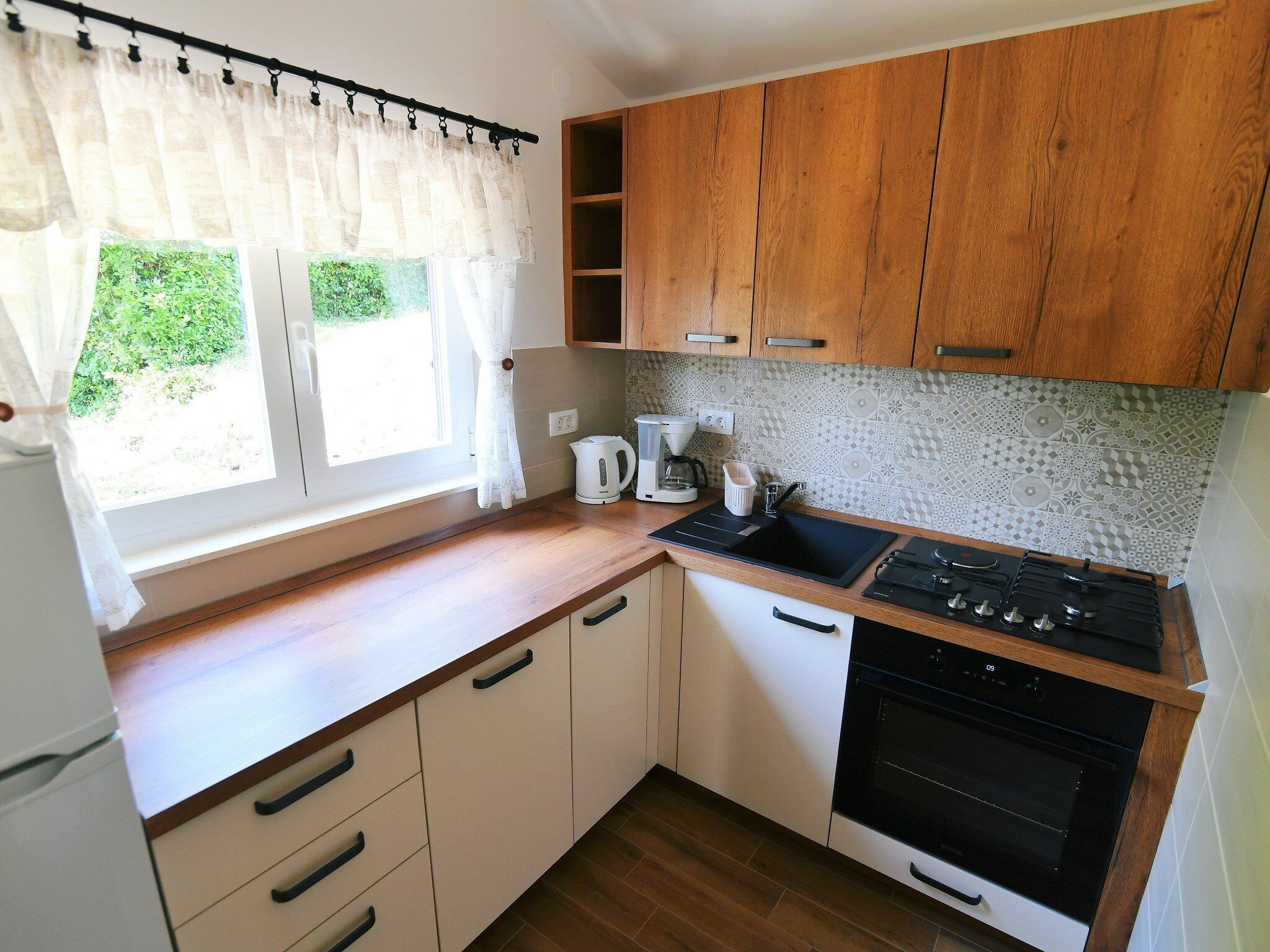 Cucina con mobili in legno, piano cottura nero, lavello e finestra con tende.