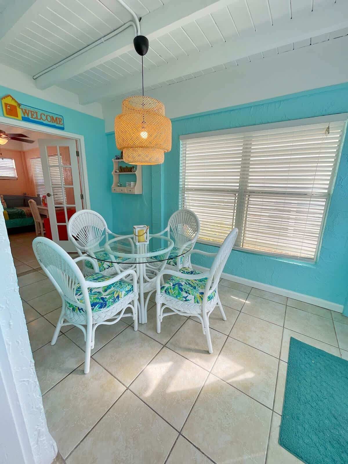 Une pièce avec un mur turquoise, une table ronde en verre entourée de chaises en osier blanc, un luminaire suspendu en fibres tressées et une fenêtre avec des stores.