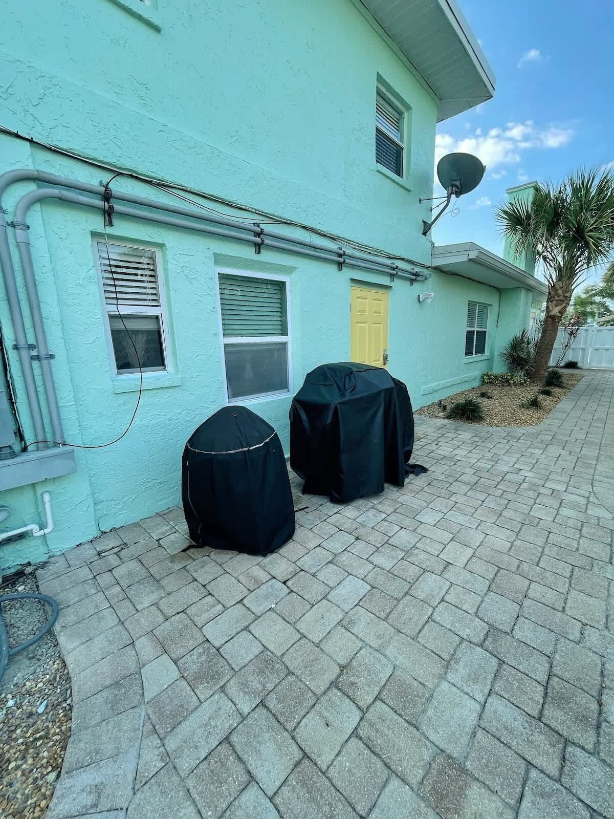 A turquoise building with a paved patio, two covered barbecue grills, and a palm tree.