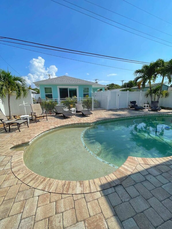 A swimming pool with a curved edge, surrounded by a paved patio with stone tiles. There are lounge chairs and a small table near the pool.