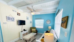 Una habitación con televisión, un banco, una mesa con lámpara, una silla y un reloj de pared.