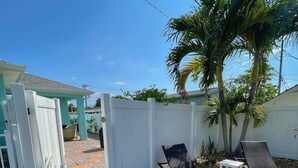 Un patio trasero con cerca blanca, dos sillones de descanso, una mesita y una palmera.