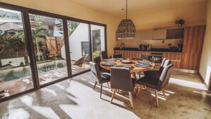 A modern dining area with a round table and chairs, a hanging light fixture, and a view of a swimming pool and garden. - Luxury Villa-4BDR Private Pool & Jacuzzi (Tulum)