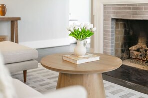 A round wooden coffee table with a white vase and book, a gray sofa, and a fireplace in the background. - Modern Villa w Pool, Hot Tub & Games! (Greensboro)