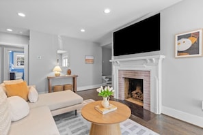 A modern living room with a sofa, a coffee table, a fireplace, and a flat-screen TV mounted above it. - Modern Villa w Pool, Hot Tub & Games! (Greensboro)