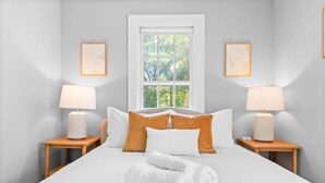Un lit bien fait, avec du linge de lit blanc, deux tables de chevet avec des lampes et une fenêtre donnant sur des arbres.