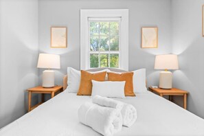A neatly made bed with white linens, two bedside tables with lamps, and a window with a view of trees.