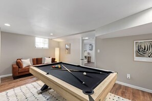 A pool table in a room with a brown sofa, a framed abstract painting, and a door leading to another room. - Modern Villa w Pool, Hot Tub & Games! (Greensboro)