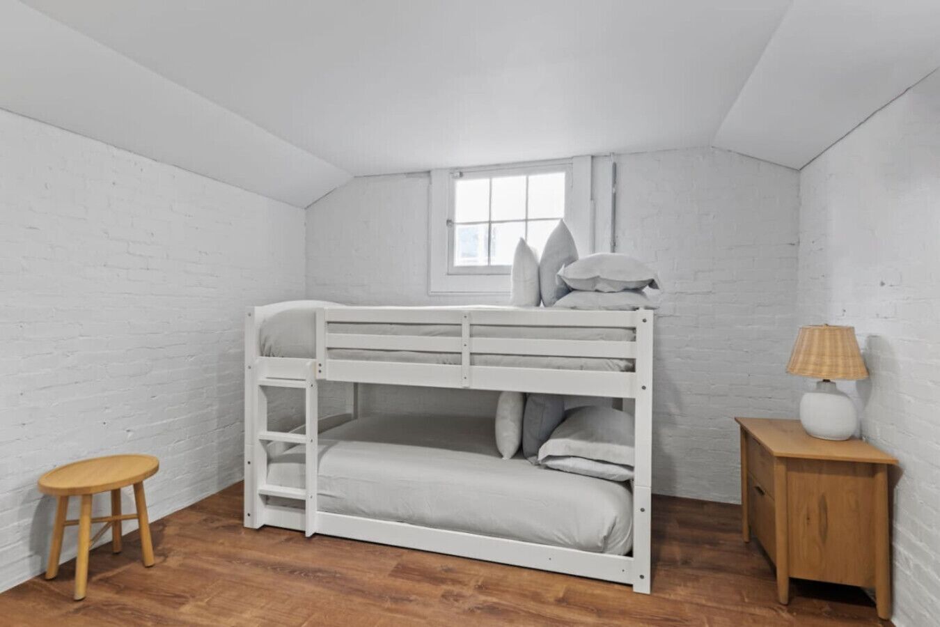 A bunk bed in a room with white brick walls, a small window, a wooden stool, and a nightstand with a lamp.