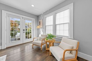 A room with a wooden floor, two white sofas, a small round table with a plant, and large windows. - Modern Villa w Pool, Hot Tub & Games! (Greensboro)