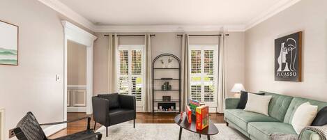 Un salon avec un canapé vert, un fauteuil noir, une table basse sur laquelle sont posés des jeux de société, une bibliothèque et un tableau accroché au mur.