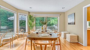 Un coin repas avec une table ronde en bois, dressée pour six personnes, entourée de six chaises. De grandes fenêtres offrent une vue sur la verdure environnante.