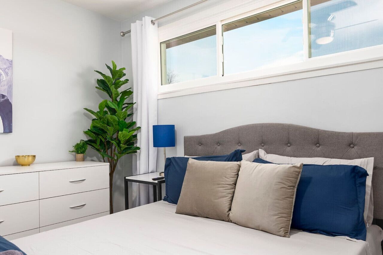 Een slaapkamer met een bed, nachtkastje, lamp, raam en een plant.