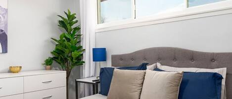 Een slaapkamer met een bed, nachtkastje, lamp, raam en een plant.