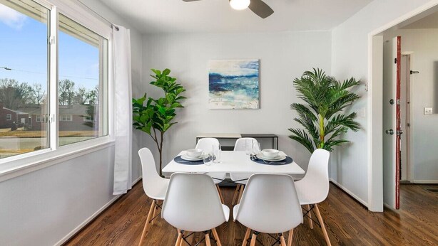 Un coin repas avec une table dressée pour quatre personnes, des chaises blanches, un ventilateur de plafond et un tableau accroché au mur.