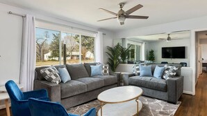 Un salon moderne avec des canapés gris, des fauteuils bleus, une table basse ronde et un ventilateur de plafond.