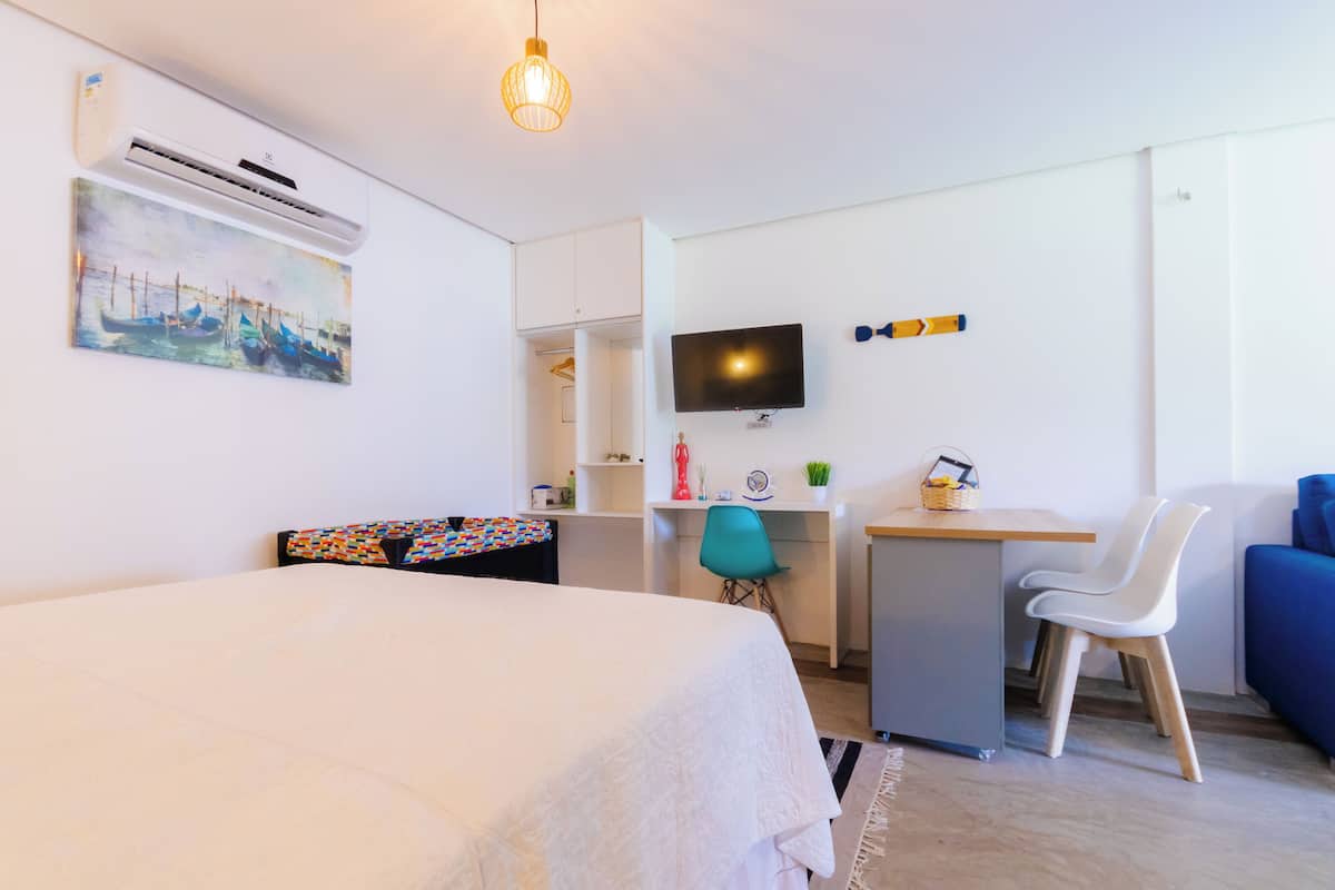A modern hotel room with a bed, desk, chair, TV, and artwork on the wall.