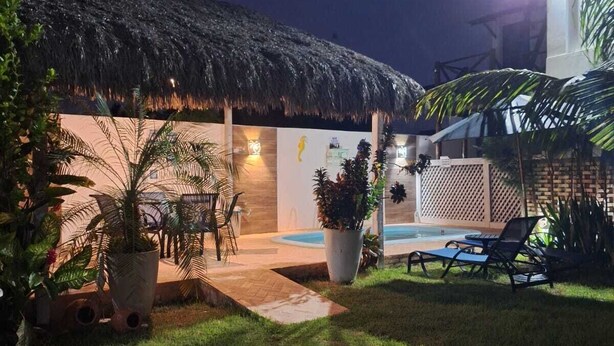 A well-lit outdoor area with a thatched-roof structure, a pool, lounge chairs, and potted plants. - Summervilleflatsbg (Cajueiro da Praia)