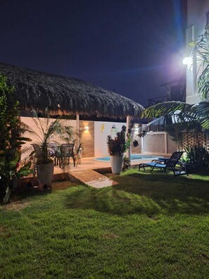 A well-lit outdoor area with a thatched-roof structure, a pool, lounge chairs, and potted plants. - Summervilleflatsbg (Cajueiro da Praia)