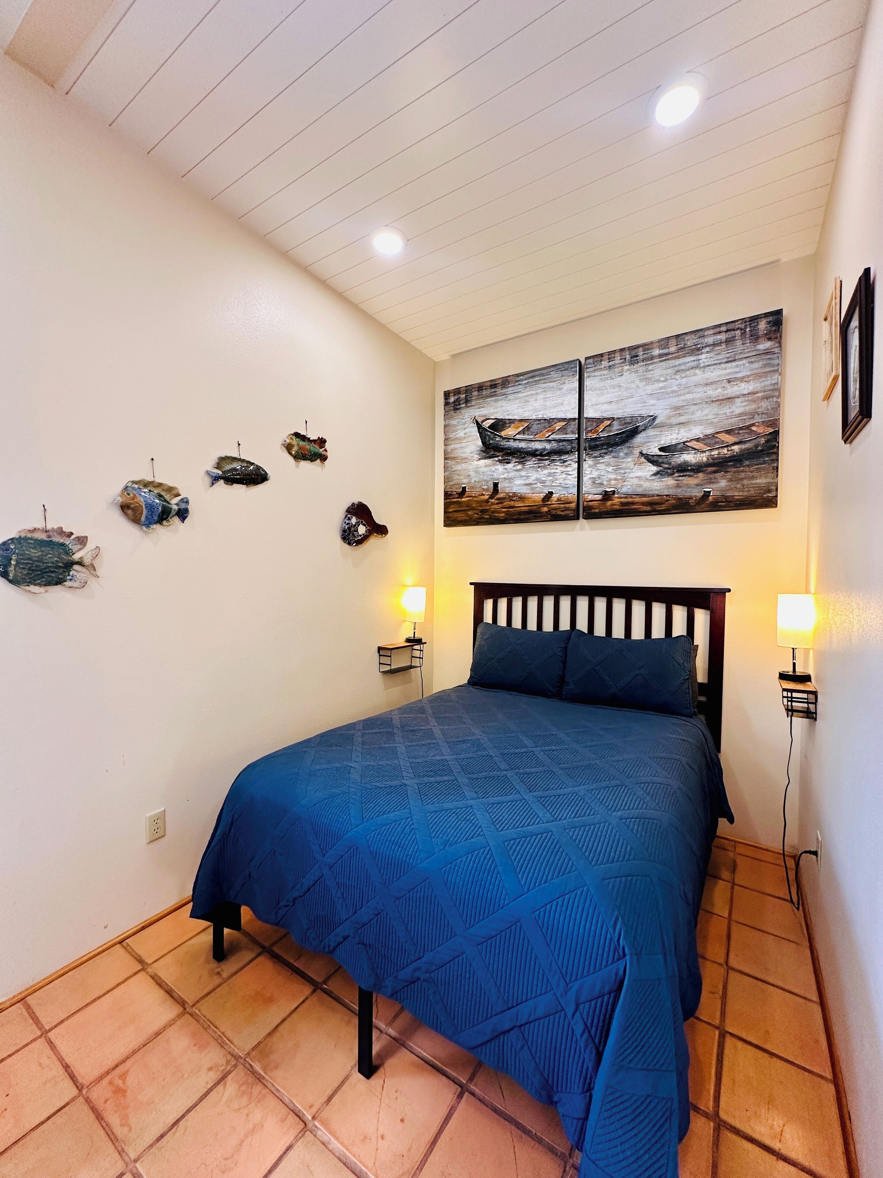 A bedroom with a bed, two wall-mounted lamps, a ceiling light, and a wall with a painting of boats.