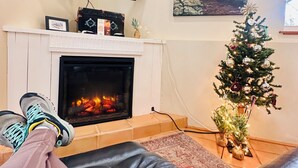 Una sala de estar con chimenea, un televisor montado encima, un árbol de Navidad y un cuadro de paisaje en la pared.