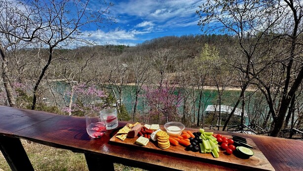 Una mesa de madera con una tabla de embutidos y bebidas, con vistas a un área boscosa con un lago.