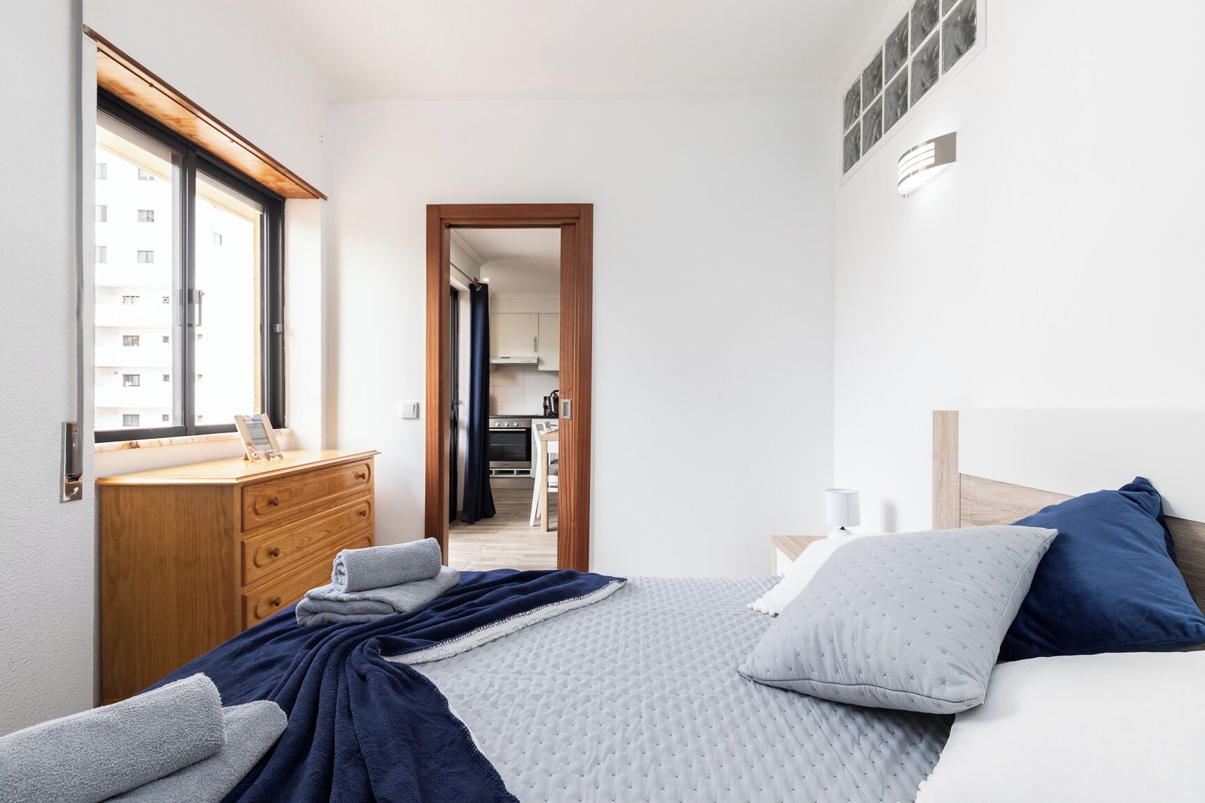 Une chambre à coucher avec un lit, une commode en bois, une fenêtre et une vue sur une cuisine et un balcon.