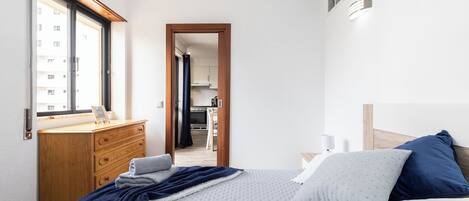 Une chambre à coucher avec un lit, une commode en bois, une fenêtre et une vue sur une cuisine et un balcon.