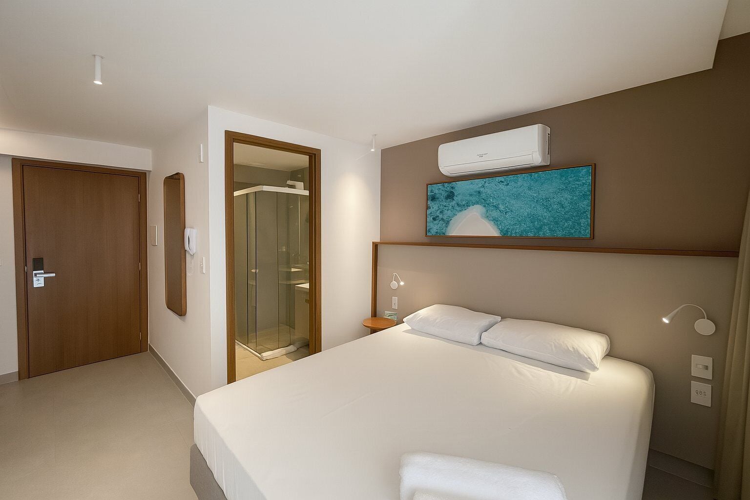 A hotel room with a bed, a shower, a mounted air conditioner, and a painting above the bed.